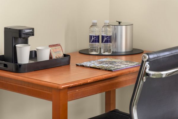 A wooden desk with a coffee maker, two cups, two water bottles, an ice bucket, and a magazine, next to a black chair.