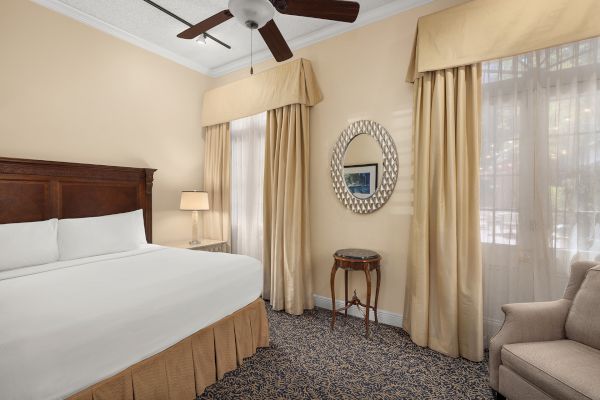 A neatly arranged bedroom with a king-sized bed, beige curtains, carpet, a mirror, a fan, a side table with a lamp, and an armchair.