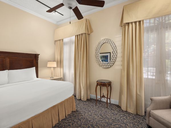 A neatly arranged bedroom with a king-sized bed, beige curtains, carpet, a mirror, a fan, a side table with a lamp, and an armchair.