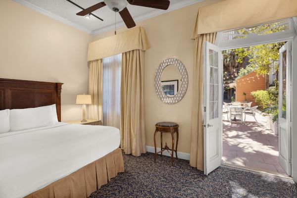 A cozy bedroom with a large bed, ceiling fan, side table, mirror, and open doors leading to a sunny patio with seating.