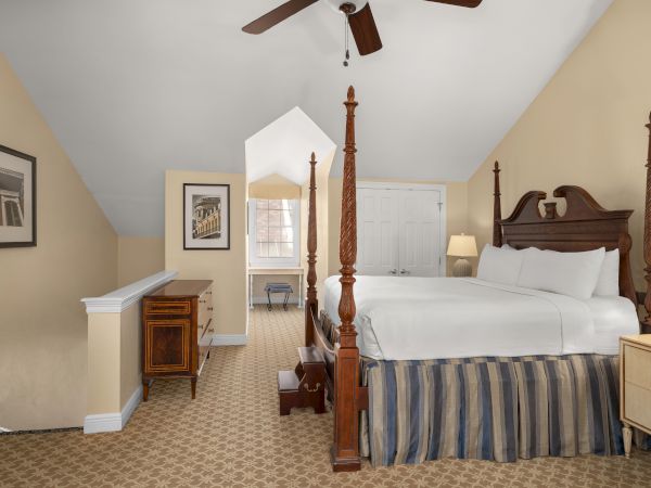 A cozy bedroom features a four-post bed, wooden furniture, beige walls, and a carpeted floor under a ceiling fan, creating a warm ambiance.