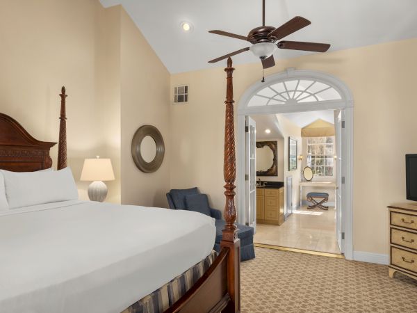 A cozy bedroom features a large bed, ceiling fan, TV, and an archway leading to a well-lit bathroom area.