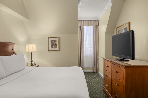 A hotel room with a bed, nightstand, lamp, wall art, window with curtains, wooden dresser, and flat-screen TV, all neatly arranged.