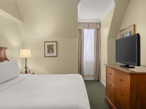 A hotel room with a bed, nightstand, lamp, wall art, window with curtains, wooden dresser, and flat-screen TV, all neatly arranged.