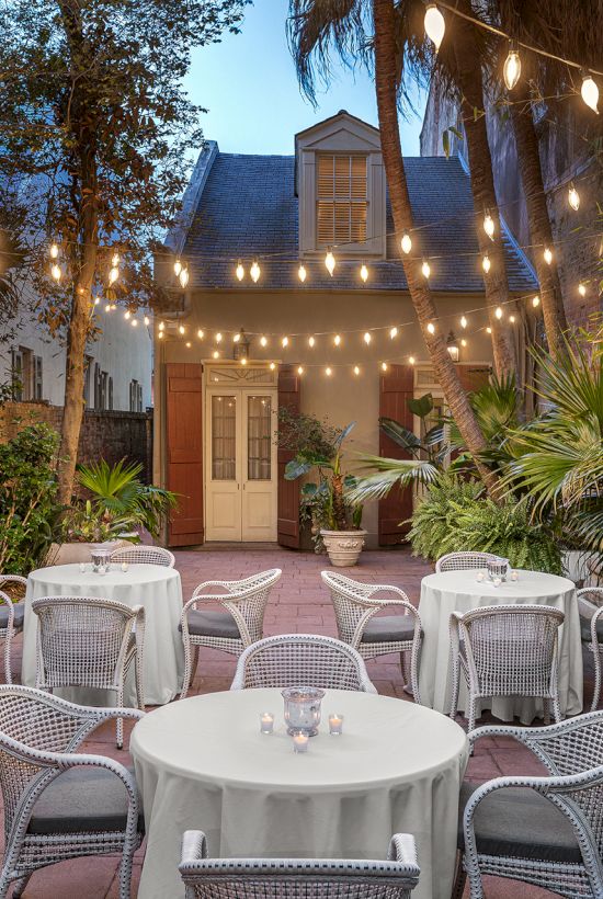 An outdoor patio with tables and chairs, surrounded by greenery, and lit by overhead string lights, set against a quaint building backdrop.