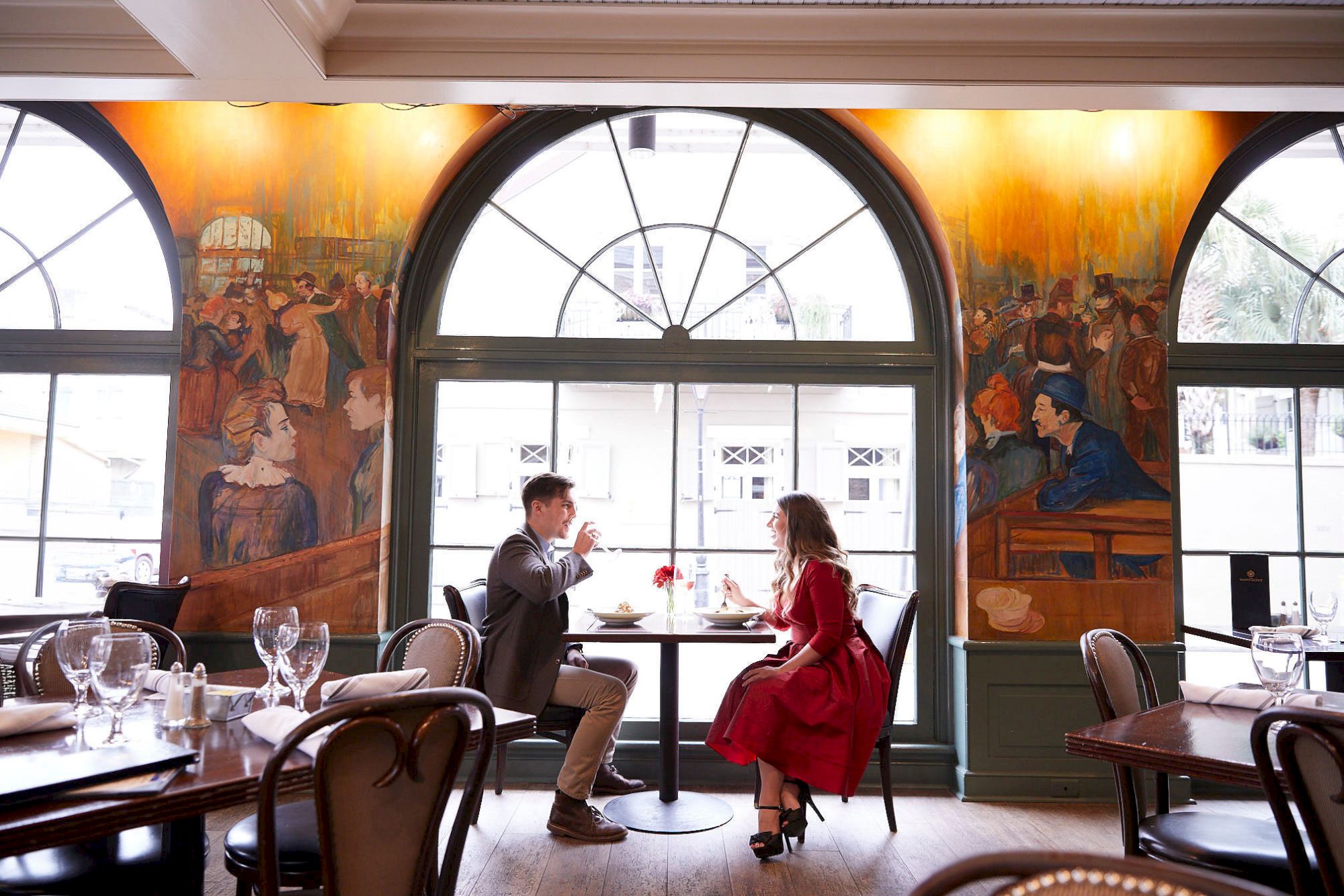 A couple dining in a restaurant with large arched windows and vibrant murals on the walls.