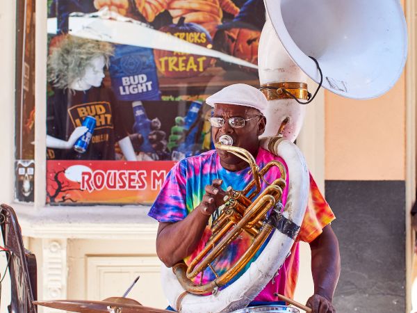 A street performer plays the sousaphone and drums simultaneously, wearing a tie-dye shirt and white cap, near a store with a window display.