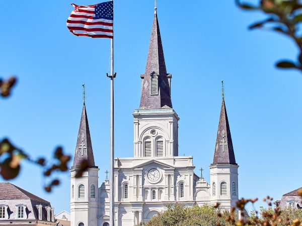A cathedral with three spires is behind a flagpole flying the American flag, framed by trees against a clear blue sky.