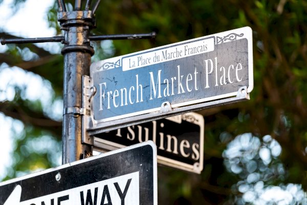 A street sign for French Market Place and Ursulines, with "One Way" below, set against a leafy background.