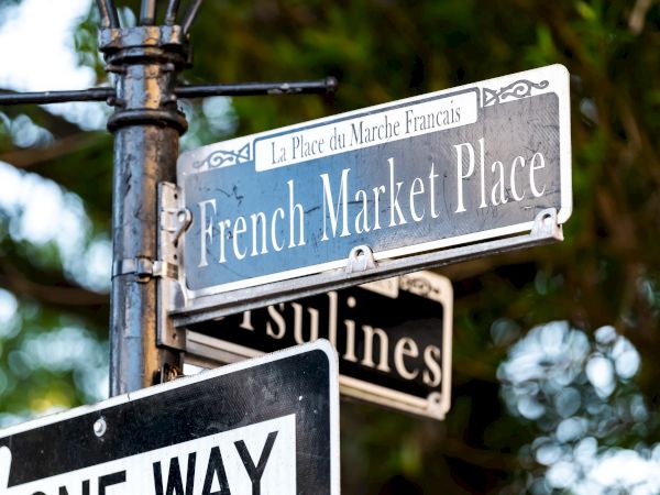 A street sign for French Market Place and Ursulines, with "One Way" below, set against a leafy background.