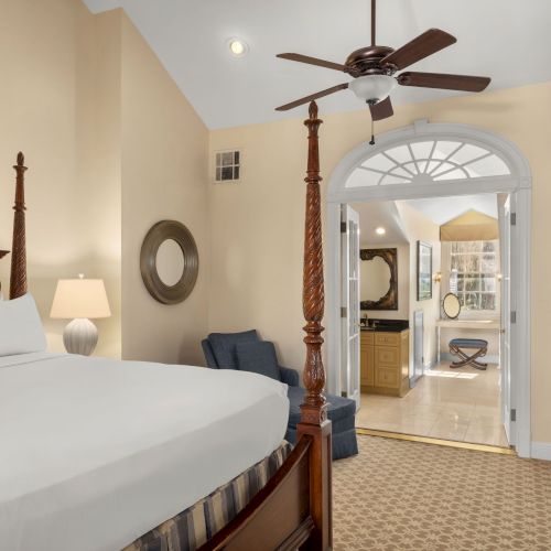 A cozy bedroom with a four-poster bed, ceiling fan, armchair, TV, dresser, and a view into a bright bathroom with vanity and stool.
