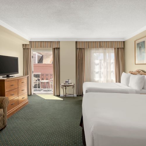 The image shows a hotel room with two beds, a TV, a dresser, a sofa, and a small table by the window leading to a balcony.