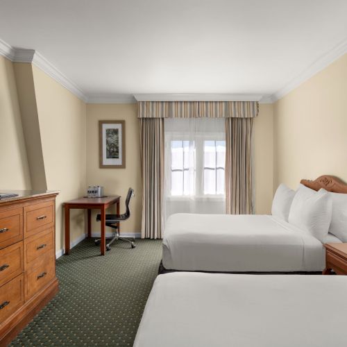 A hotel room with two beds, TV, desk, chair, and window with curtains. There’s also a nightstand with a lamp and a framed picture on the wall.