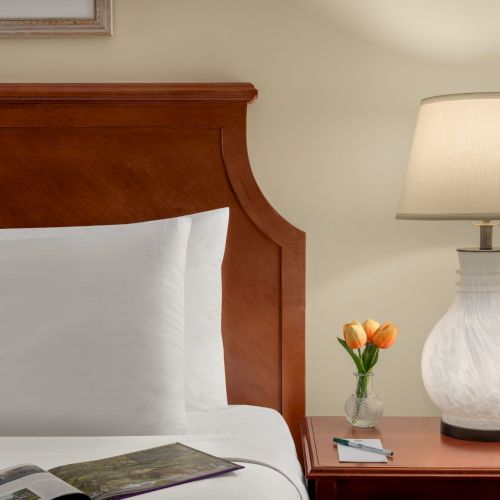A bed with white pillows, a magazine, wooden headboard, beside a nightstand with a lit lamp, vase with tulips, and a pen on paper.