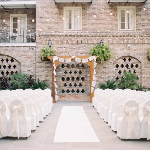 An outdoor wedding setup with white covered chairs, a wooden arch with decorations, a white aisle runner, and brick wall backdrop.