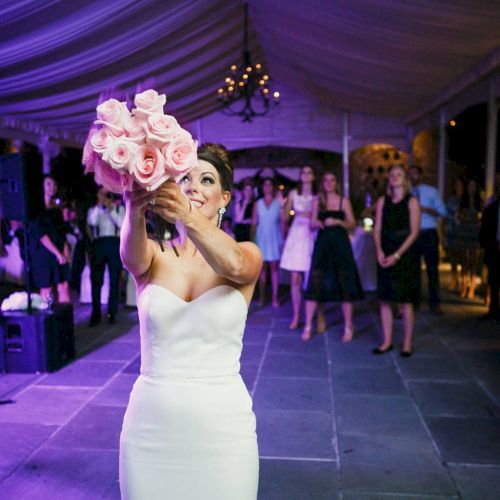 A bride is joyfully tossing a bouquet of pink roses at a wedding reception while guests eagerly watch.