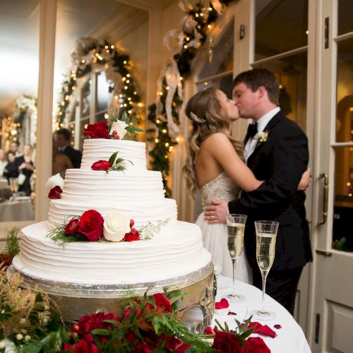 A couple kisses at a wedding, with a tiered cake decorated with red and white roses in the foreground, champagne glasses nearby.