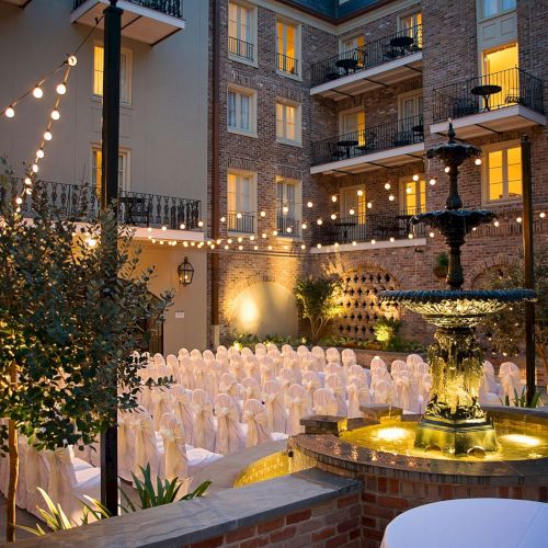 Outdoor event setup with chairs covered in white fabric, string lights overhead, and a lit fountain in a courtyard surrounded by buildings.