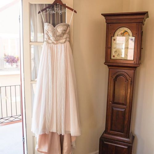 A wedding dress hangs on a door beside a wooden grandfather clock in a room with textured walls and carpet.