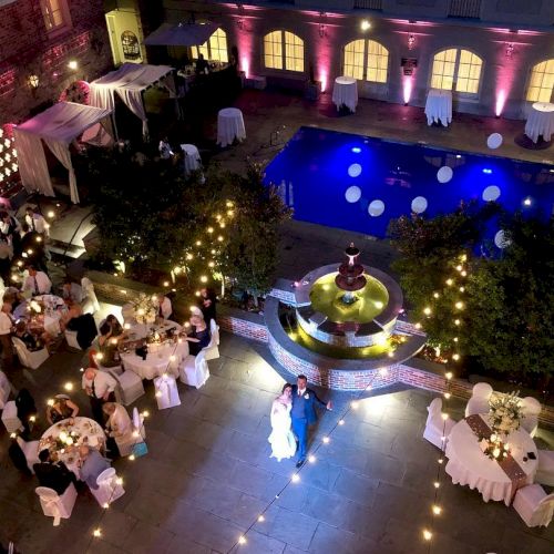 A nighttime event with tables, string lights, and a pool surrounded by a courtyard. There's a fountain and a couple in the center.