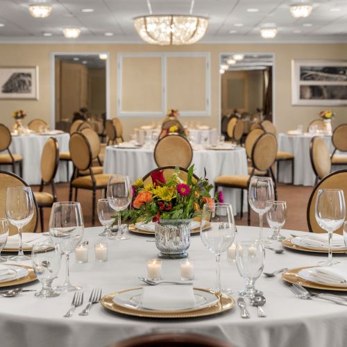 Elegant banquet hall with round tables, table settings, floral centerpieces, chandeliers, mirrors, and artwork on beige walls.