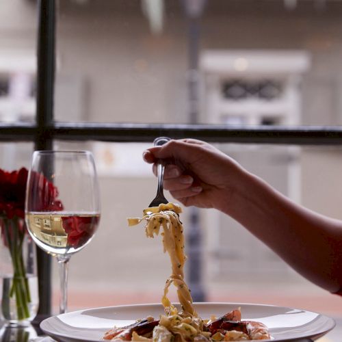 A hand twirls pasta with a fork above a plate, next to a glass of white wine and red flowers by a window.