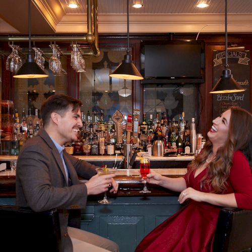 A couple laughs and enjoys drinks at a lively bar, surrounded by a variety of bottles and stylish decor.
