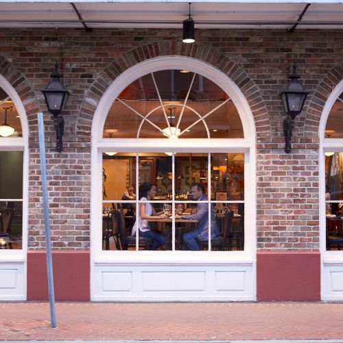 A brick-facade restaurant with large arched windows shows a couple sitting inside, enjoying a meal together.