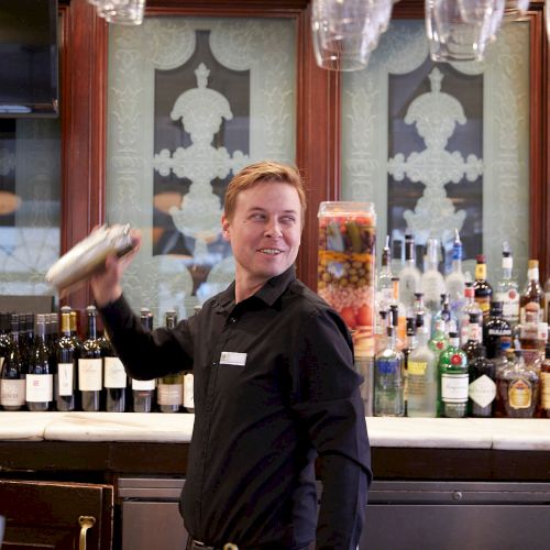 A bartender is shaking a cocktail shaker behind a bar with liquor bottles and a glass of martini in the foreground.