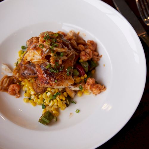 A plate with seasoned chicken over corn, shrimp, and vegetables, garnished with chopped green onions.