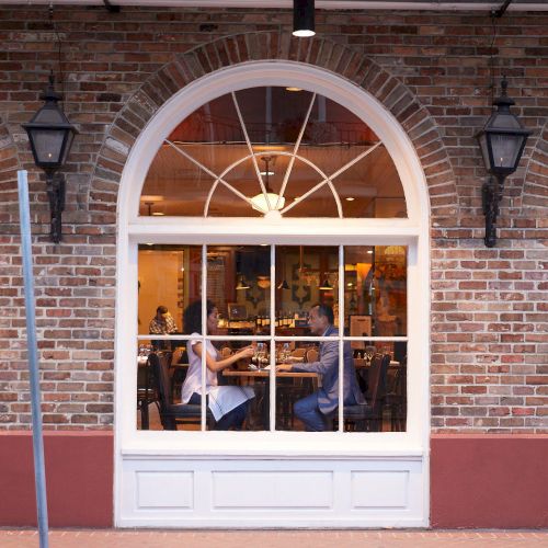 A couple dines inside a cozy, brick-walled restaurant, visible through a large arched window with warm interior lighting.