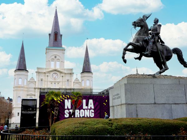 A church with three spires, a statue of a rider on a horse, and a "NOLA STRONG" sign are in the image.