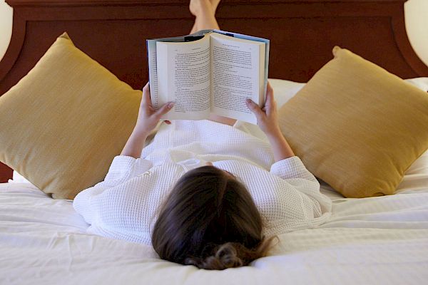 A person in a white robe is lying back on a bed, reading a book, with legs propped up on a headboard.