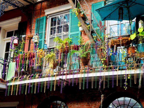 A colorful balcony with plants and vibrant beads, set against a building with turquoise shutters and exposed brick.