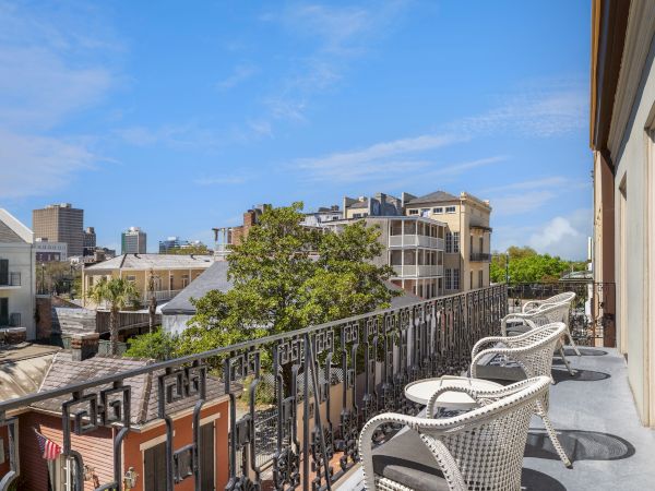 A balcony with wicker chairs overlooks an urban scene, featuring several buildings and a tree under a clear blue sky.