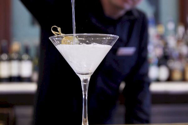 A bartender is pouring a clear liquid into a martini glass on a counter, with a garnish on the rim.