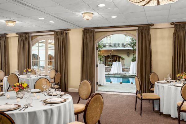 Elegant indoor dining setup with round tables by a poolside courtyard, featuring white tablecloths and floral centerpieces.