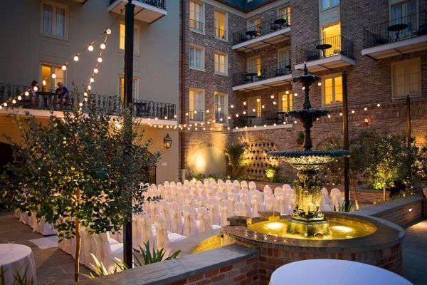 An elegant courtyard is set up for an event with chairs, a fountain, string lights, and a brick building backdrop, creating a cozy ambiance.