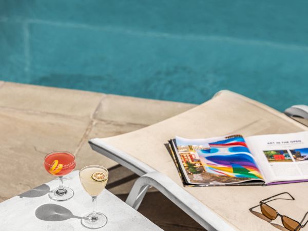 Two cocktails on a table, an open magazine, sunglasses, and a poolside lounge chair by a swimming pool.