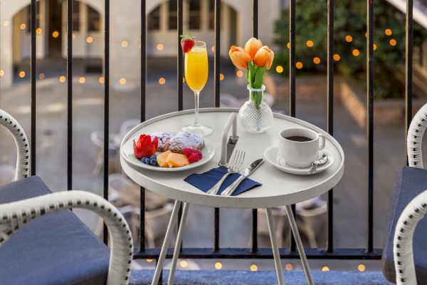 A small table on a balcony with a breakfast spread: coffee, a plate of fruit, a mimosa, silverware on a napkin, and a vase with tulips.