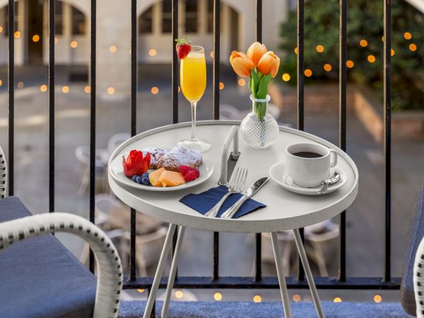 A small table on a balcony with a breakfast spread: coffee, a plate of fruit, a mimosa, silverware on a napkin, and a vase with tulips.