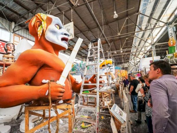 A craftsman workshop with large orange mascot statues under construction, ladders and tools, workers and onlookers in a busy market-like space.