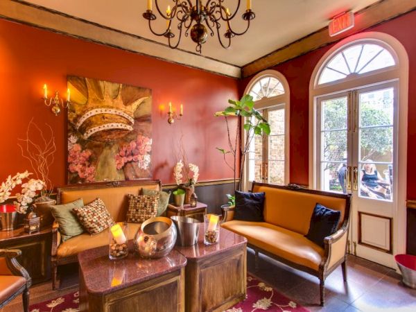 A cozy, warmly lit lounge with red walls, sofas, plants, and a central coffee table, near arched windows and a chandelier.