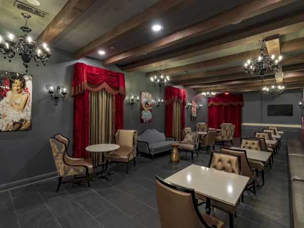 A chic lounge with dark walls, red drapes, chandeliers, and stylish seating; small tables and modern booths create an intimate, club-like vibe.
