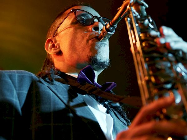 A saxophonist in a suit and sunglasses plays passionately into a microphone, under warm stage lights, caught in a live performance.