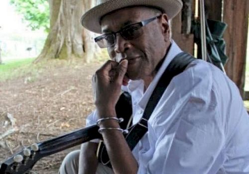 A man wearing a hat and sunglasses sits with a guitar, resting his hand on his chin under a shaded outdoor setting, looking thoughtful.