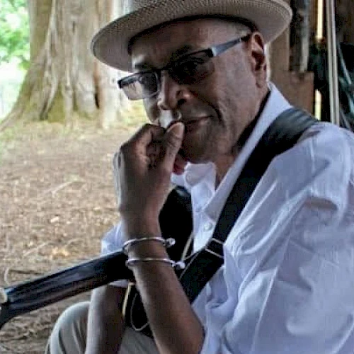 A man wearing a hat and sunglasses sits with a guitar, resting his hand on his chin under a shaded outdoor setting, looking thoughtful.