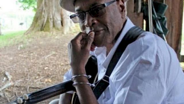 A man wearing a hat and sunglasses sits with a guitar, resting his hand on his chin under a shaded outdoor setting, looking thoughtful.