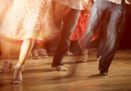 People dancing in a warmly lit scene, showing legs and feet in motion on a wooden floor, wearing colorful skirts and pants, mid-dance.