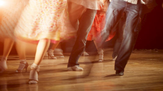 People dancing in a warmly lit scene, showing legs and feet in motion on a wooden floor, wearing colorful skirts and pants, mid-dance.